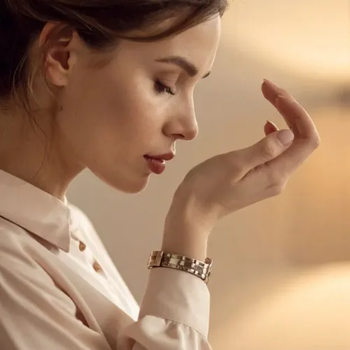 Close-up profile of a woman examining her hand and nails, wearing a beige blouse and a metal watch in warm indoor lighting.