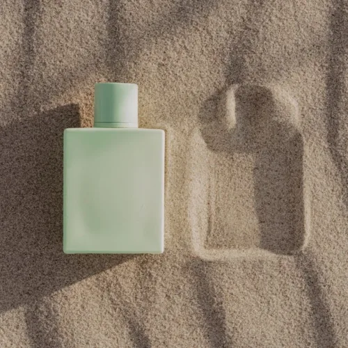 Mint-green rectangular bottle on beach sand with a square imprint nearby.