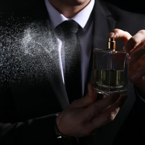 Man in a suit sprays perfume from a square glass bottle, mist billowing to the left.
