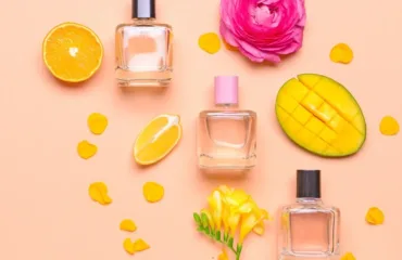 Flat lay of three perfume bottles surrounded by a sliced orange, mango half, pink rose, yellow flower, and scattered petals on a peach background.