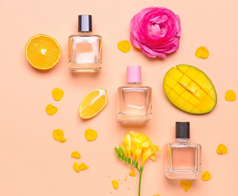 Flat lay of three perfume bottles surrounded by a sliced orange, mango half, pink rose, yellow flower, and scattered petals on a peach background.