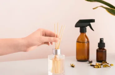 Hand inserts diffuser reeds into a square glass bottle of scented oil on a white table.