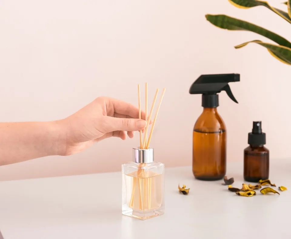 Hand inserts diffuser reeds into a square glass bottle of scented oil on a white table.