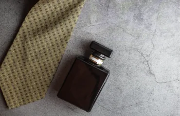 Naykrashchi parfumy dlya cholovikiv u podarunok universalna dobirka Dark brown square perfume bottle with a black cap lying next to a tan dotted tie on a gray concrete surface, forming a stylish flat lay.