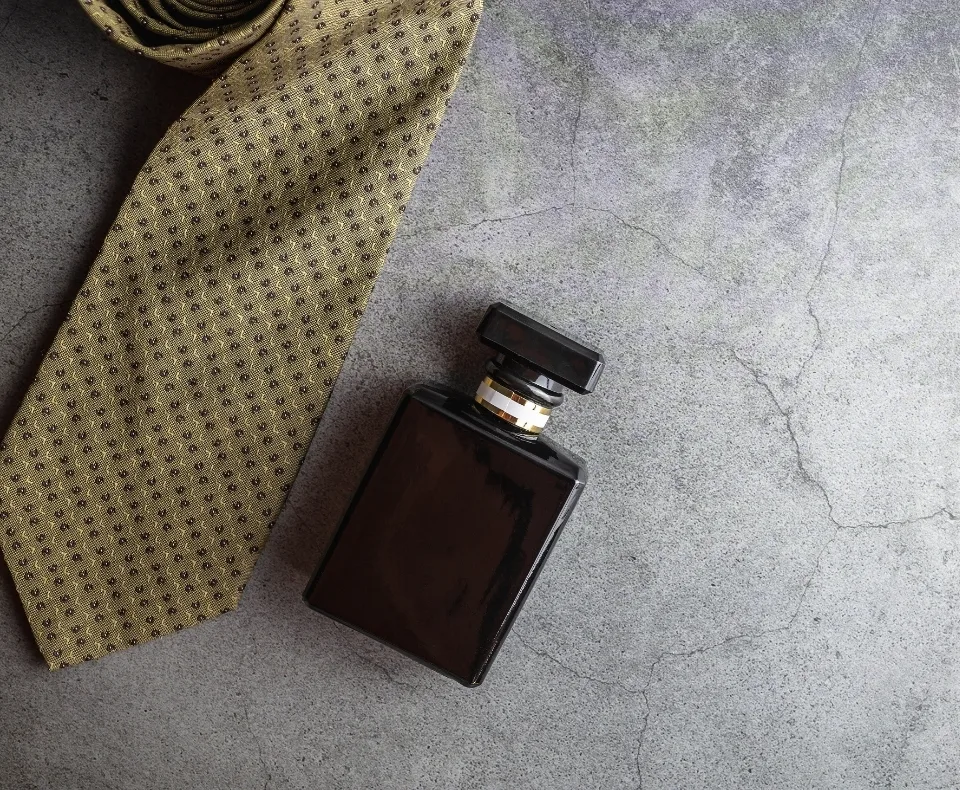 Dark brown square perfume bottle with a black cap lying next to a tan dotted tie on a gray concrete surface, forming a stylish flat lay.
