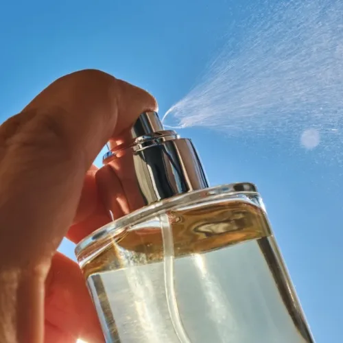 Close-up of a hand pressing a perfume spray bottle outdoors, mist spraying into a clear blue sky.