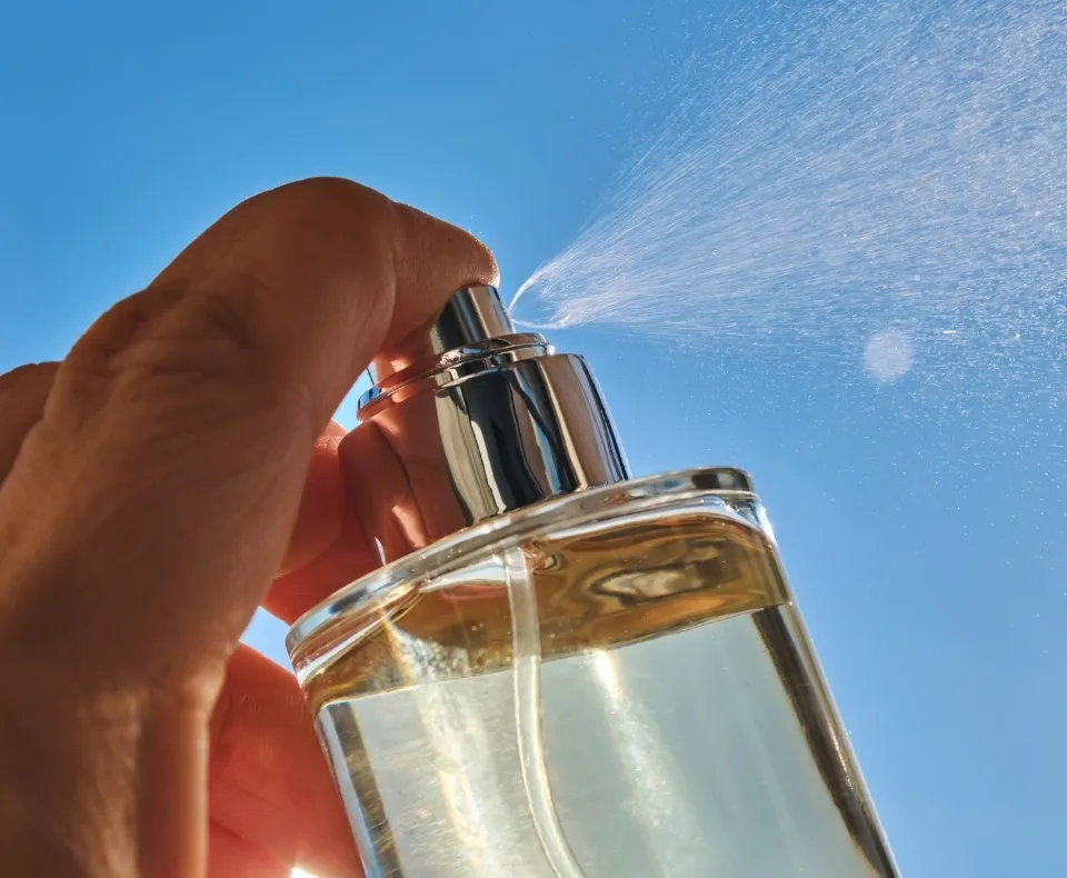 Close-up of a hand pressing a perfume spray bottle outdoors, mist spraying into a clear blue sky.